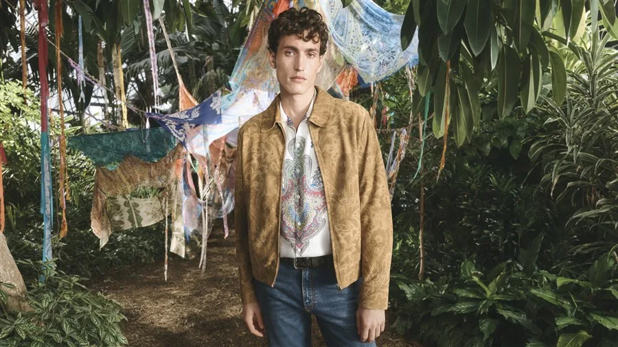 A young man in a tan paisley jacket and printed shirt stands amid lush tropical foliage and colorful draped fabrics.