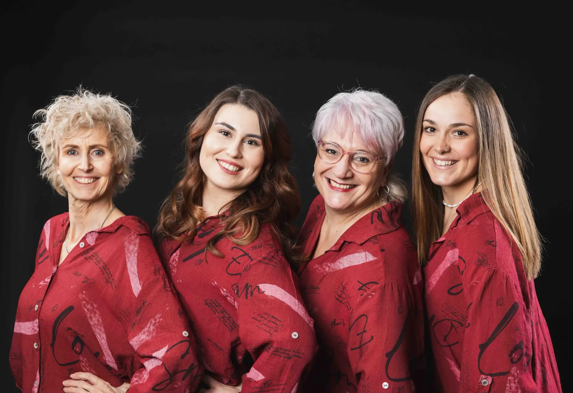 Four women in matching red patterned shirts pose together smiling warmly in a professional studio setting.