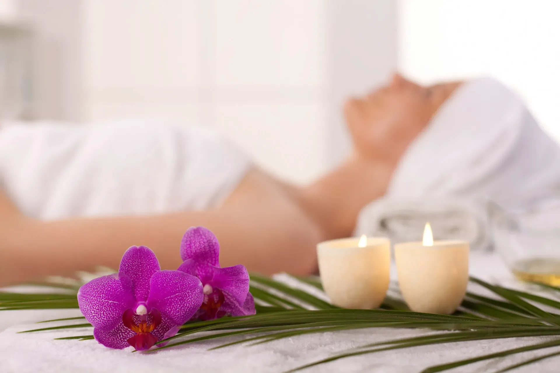 A client relaxes on a treatment bed surrounded by orchids and glowing candles in a serene spa setting.