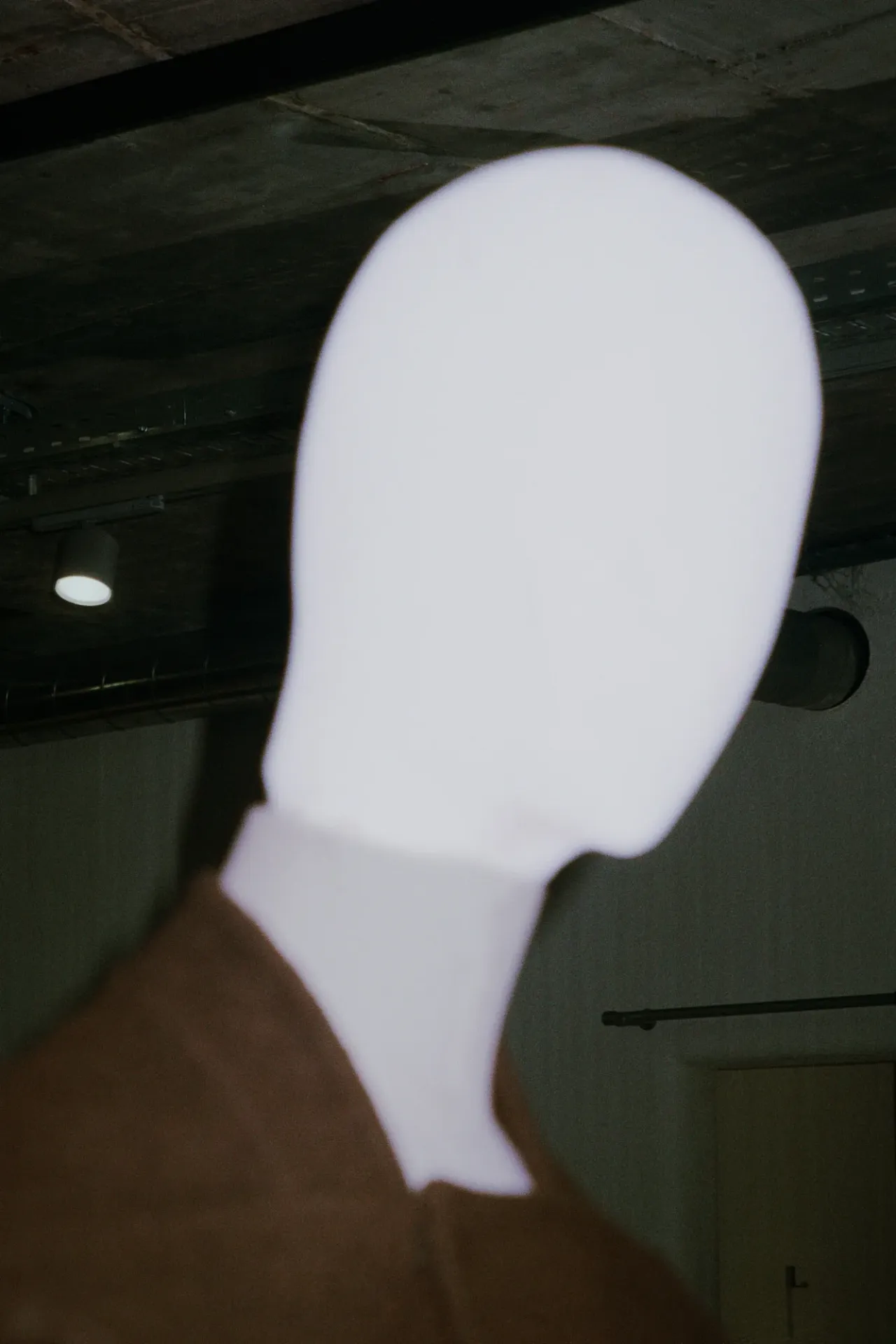 Blurred silhouette of a figure with an overexposed white head against a dark industrial ceiling space.